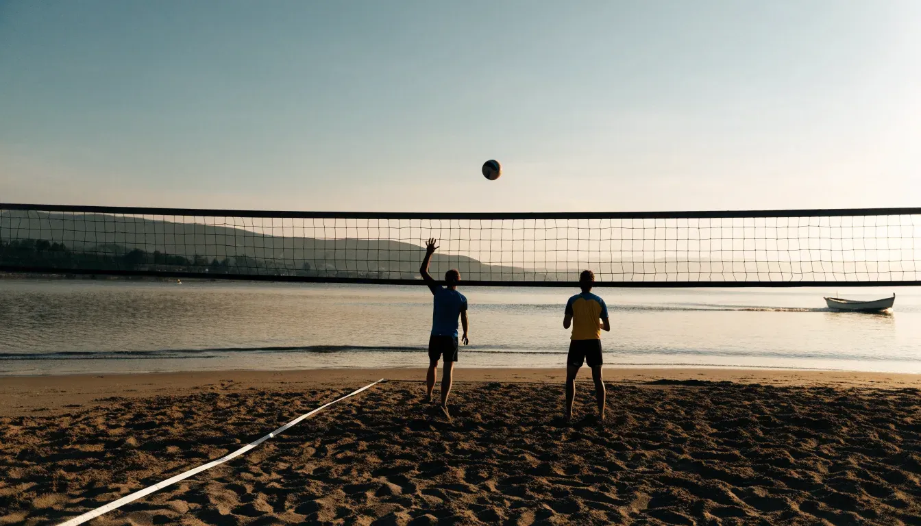 Ein Volleyball liegt im Sand eines Spielfelds am Seeufer, im Hintergrund gleitet ein Boot über das Wasser.