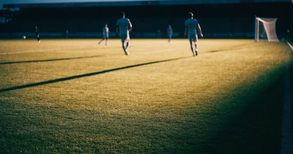 Ein Fussballplatz im Abendlicht mit langen Schatten der Torpfosten, die Rasenfläche leuchtet gelblich.