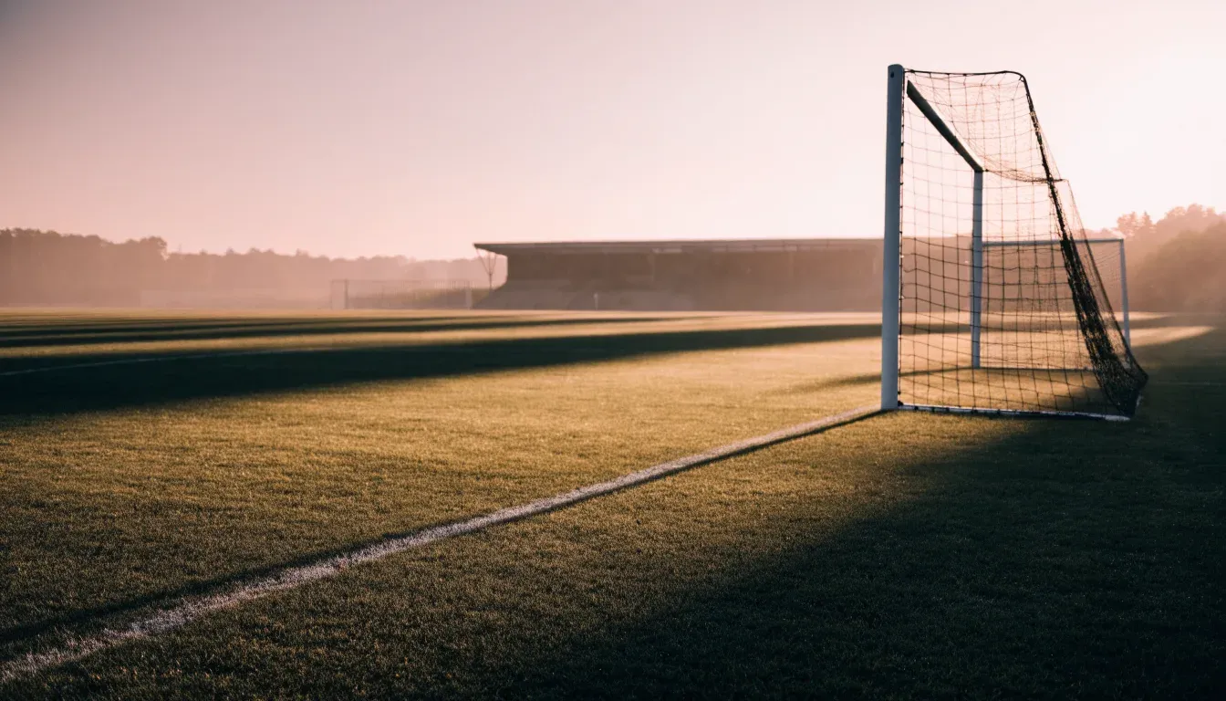 Ein Fussballtor mit Netz in der Totalen auf einem leeren Platz, Tau glänzt im Gras und die Morgensonne steht tief.