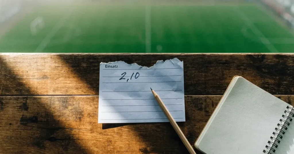 Ein Klemmbrett mit leerem Papier und ein Bleistift liegen auf einer Tribünenbank, darunter ein Rasenplatz im Frühnebel.