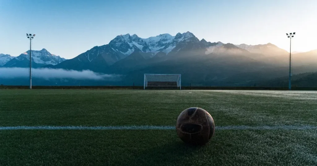 Ein Fussball liegt am frühen Morgen auf dem Rasen eines leeren Fussballfeldes, dahinter eine kleine Tribüne und schneebedeckte Berge.