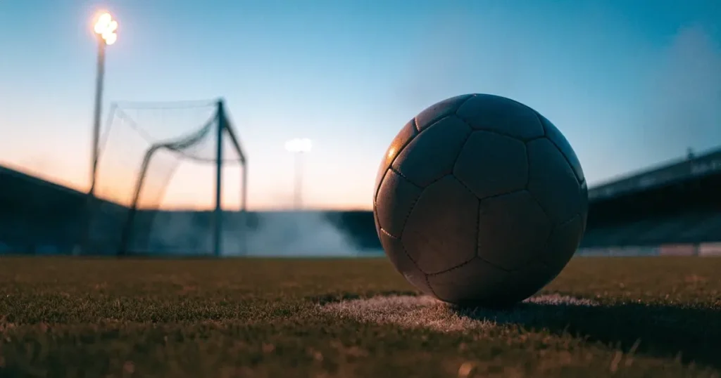 Ein Fussball liegt auf dem Anstosspunkt eines leeren Rasenplatzes, im Hintergrund glimmen Flutlichtmasten im Abendlicht.