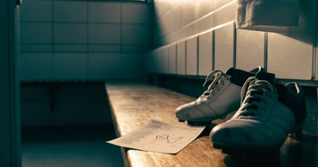 Ein Paar Fussballschuhe steht auf einer Holzbank in einer leeren Umkleide, über der Bank hängt ein Handtuch.